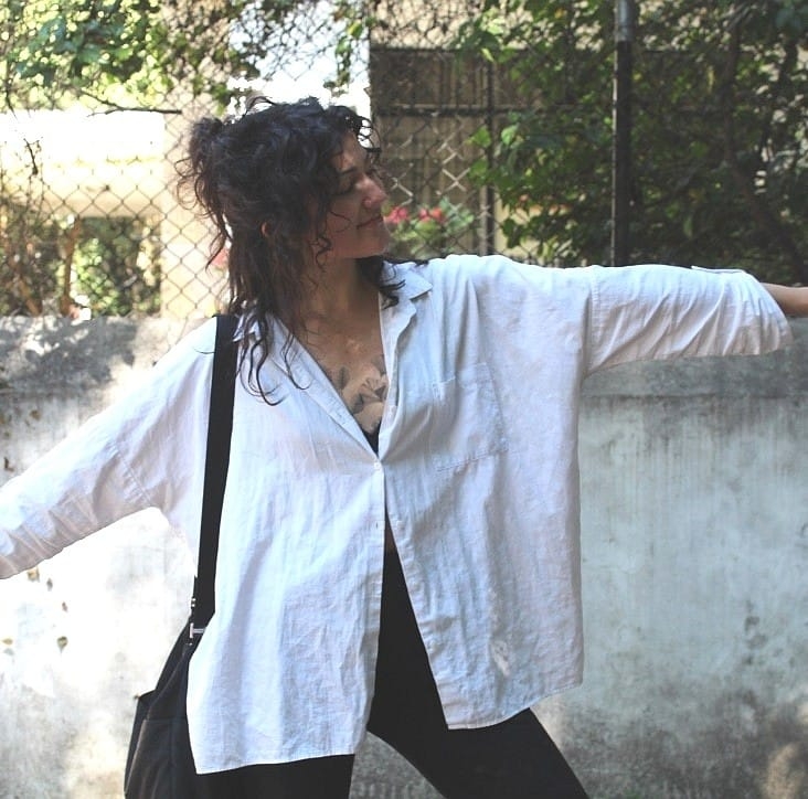 Pilar Waters mid-movement in an oversized white shirt against a weathered grey wall, arm reaching out to the right.