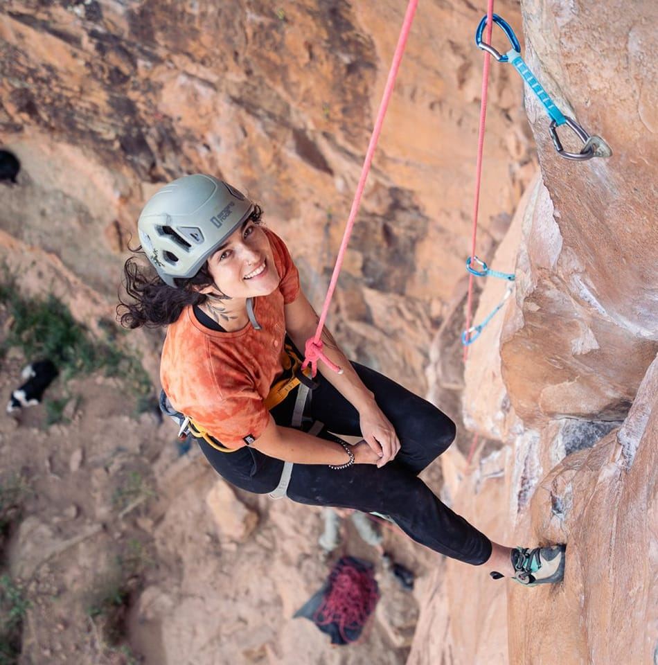 Pilar Waters in a climbing helmet on a sandstone wall, smiling up at the camera with blue carabiners and a rope in frame.