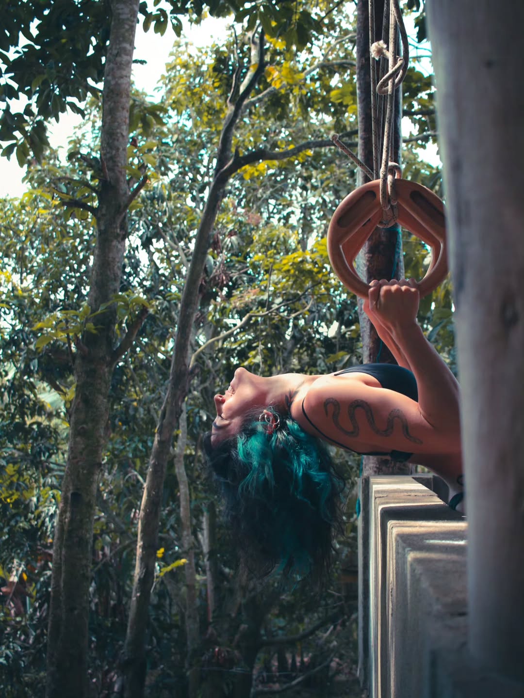 Pilar Waters hanging horizontally from gymnastic rings tied to jungle trees, head back, holding the rings overhead with one arm.