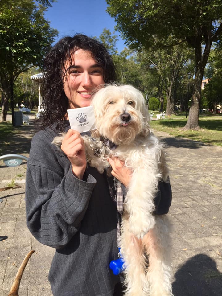Pilar Waters smiling outdoors in a park, holding a small light-haired dog in her arms.